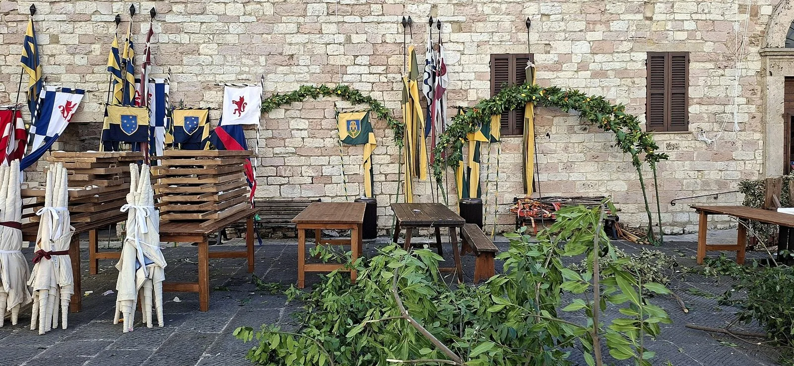 Stendardi e bandiere colorate appoggiati a un muro in pietra ad Assisi, con archi di fronde e tavoli in legno allestiti per il Calendimaggio