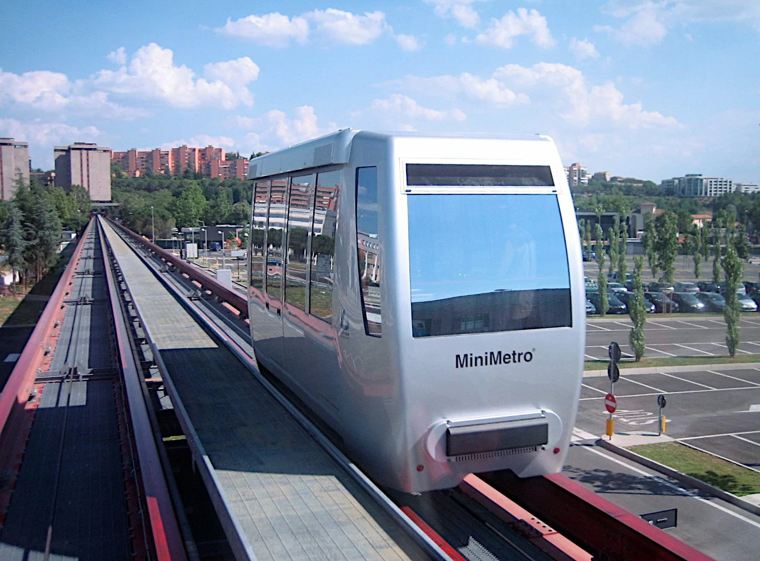 Una cabina argentata del minimetrò di Perugia sulla via di corsa sopraelevata, con un ampio parcheggio sulla destra